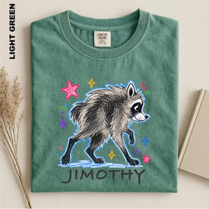 May include: A light green t-shirt featuring a charcoal-colored sketch of a raccoon walking. The design includes colorful hand-drawn stars and sparkles in pink, yellow, and blue. The name JIMOTHY is printed in dark gray letters below the raccoon.