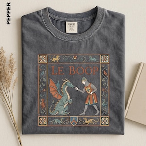 May include: A charcoal gray crew neck t-shirt featuring a medieval-style graphic print. The design shows a knight touching a dragon's nose with the text LE BOOP, surrounded by a decorative floral border and small mythical creatures.