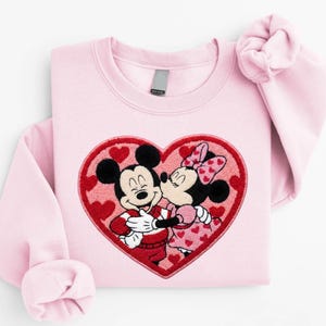 May include: Light pink sweatshirt featuring an embroidered design of Mickey Mouse and Minnie Mouse embracing within a red heart. The design includes red hearts and the text "SunsetAndStitchCo" and "Light Pink".