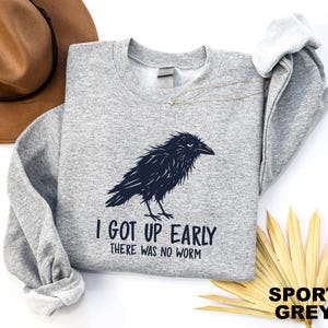 May include: A sport grey crewneck sweatshirt featuring a black graphic of a ruffled crow. Below the bird, the text reads "I GOT UP EARLY" and "THERE WAS NO WORM" in a dark, sans-serif font. The garment has long sleeves and a ribbed neckline.