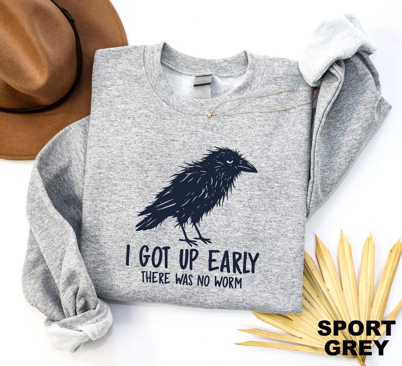 May include: A sport grey crewneck sweatshirt featuring a black graphic of a ruffled crow. Below the bird, the text reads "I GOT UP EARLY" and "THERE WAS NO WORM" in a dark, sans-serif font. The garment has long sleeves and a ribbed neckline.