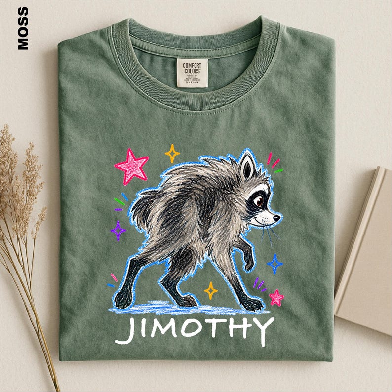 May include: A moss green crew neck t-shirt featuring a graphic of a gray raccoon with a sketch-like texture. The design includes colorful stars and the text "JIMOTHY" in white lettering below the raccoon. The shirt has a Comfort Colors label.