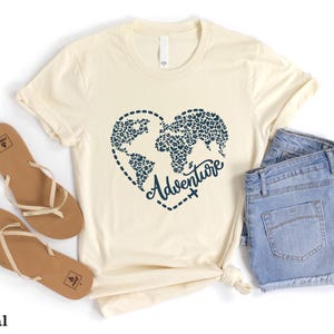 May include: Cream-colored t-shirt with a navy blue heart-shaped world map design and the word "Adventure". The shirt is paired with blue denim shorts and brown sandals. The text "SunsetAndStitchCo" is visible.