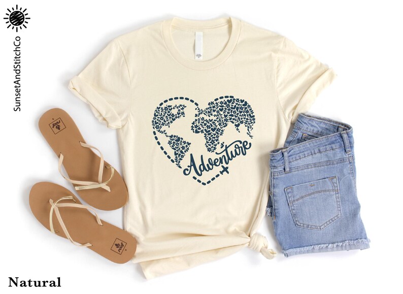May include: Cream-colored t-shirt with a navy blue heart-shaped world map design and the word "Adventure". The shirt is paired with blue denim shorts and brown sandals. The text "SunsetAndStitchCo" is visible.