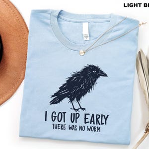 May include: A light blue crew neck t-shirt featuring a dark navy graphic of a ruffled crow. Below the bird, the text reads, "I GOT UP EARLY THERE WAS NO WORM" in a bold, dark navy font. The shirt has a white interior neck label.