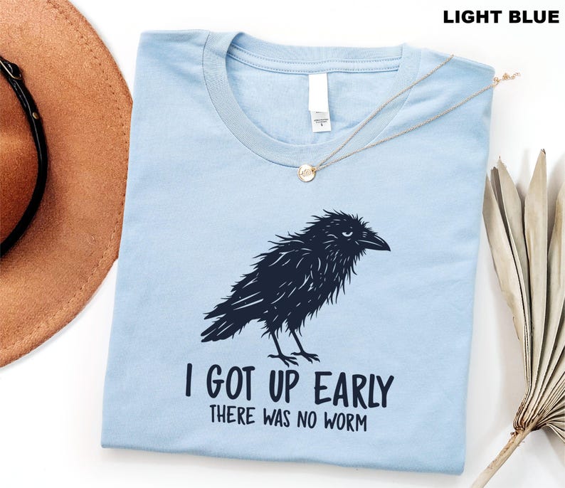 May include: A light blue crew neck t-shirt featuring a dark navy graphic of a ruffled crow. Below the bird, the text reads, "I GOT UP EARLY THERE WAS NO WORM" in a bold, dark navy font. The shirt has a white interior neck label.