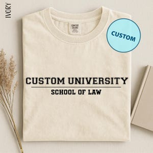May include: Ivory-colored t-shirt with the text "CUSTOM UNIVERSITY SCHOOL OF LAW" in black. A light blue circle with the word "CUSTOM" is in the upper right corner. The shirt is folded and placed on a light-colored surface.