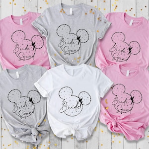 May include: A collection of t-shirts in pink, gray, and white, each featuring a black Mickey Mouse-shaped design filled with stars and the words "Bride Squad" or "Bride". The shirts are short-sleeved.