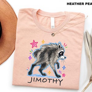 May include: A heather peach t-shirt featuring a graphic illustration of a grey and black raccoon walking. The design includes colorful hand-drawn style stars and the name JIMOTHY printed in black capital letters below the raccoon character.