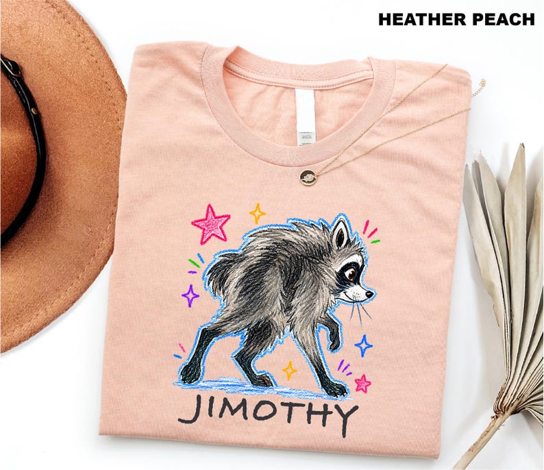 May include: A heather peach t-shirt featuring a graphic illustration of a grey and black raccoon walking. The design includes colorful hand-drawn style stars and the name JIMOTHY printed in black capital letters below the raccoon character.