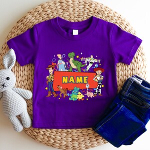 A purple children's t-shirt with a character design and the word "NAME" printed on it, along with a crocheted rabbit toy and a pair of blue jeans.