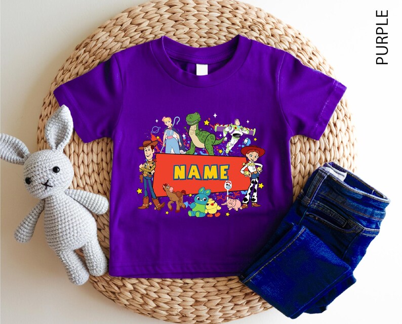 A purple children's t-shirt with a character design and the word "NAME" printed on it, along with a crocheted rabbit toy and a pair of blue jeans.