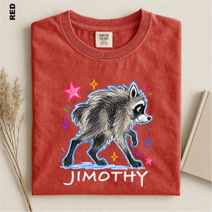 May include: A red t-shirt featuring a graphic illustration of a gray raccoon with black markings. The raccoon is surrounded by colorful, hand-drawn style stars and dashes. The name JIMOTHY is printed in white capital letters below the raccoon.