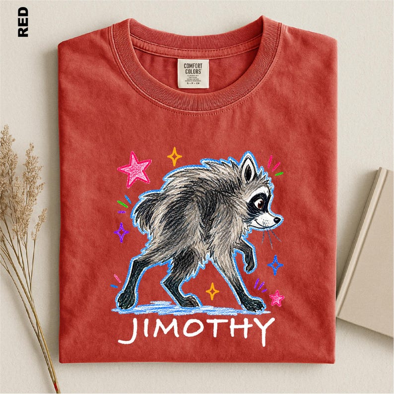 May include: A red t-shirt featuring a graphic illustration of a gray raccoon with black markings. The raccoon is surrounded by colorful, hand-drawn style stars and dashes. The name JIMOTHY is printed in white capital letters below the raccoon.