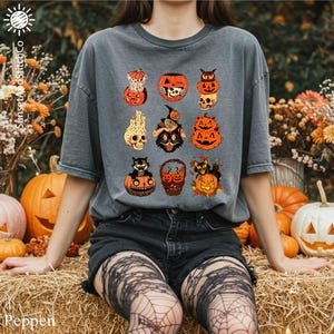 A person wearing a gray t-shirt with a Halloween-themed graphic of various spooky and festive characters, including jack-o-lanterns, bats, and skulls, is sitting on hay bales surrounded by pumpkins and flowers.