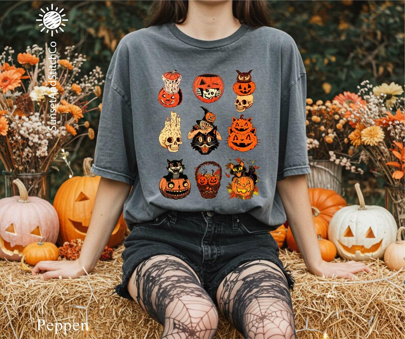A person wearing a gray t-shirt with a Halloween-themed graphic of various spooky and festive characters, including jack-o-lanterns, bats, and skulls, is sitting on hay bales surrounded by pumpkins and flowers.
