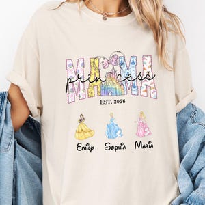May include: Ivory-colored t-shirt with the words "MAMA princess" in a colorful design, featuring a castle and three princess illustrations. Below the princesses are the names Emily, Sophia, and Maria. The shirt is a casual style.