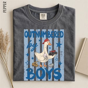 A gray t-shirt with a graphic design featuring a cartoon duck holding a baseball bat and a truck, along with the text "OUTNumbered by BOYS".