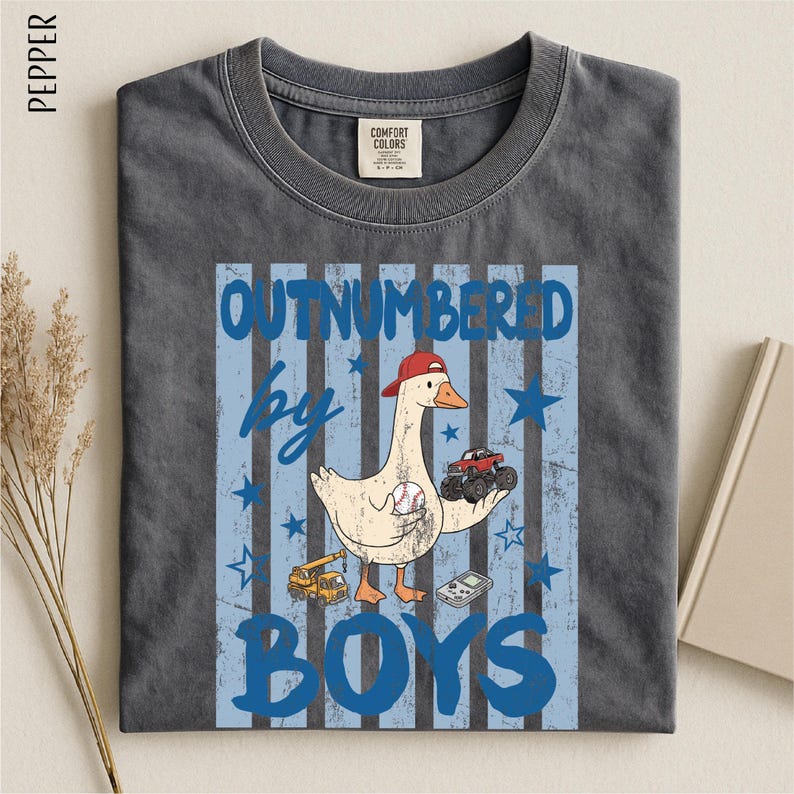 A gray t-shirt with a graphic design featuring a cartoon duck holding a baseball bat and a truck, along with the text "OUTNumbered by BOYS".