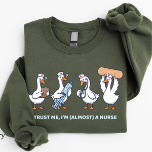May include: Military green sweatshirt featuring a humorous graphic of four cartoon geese dressed as nurses, each holding medical tools. The text "TRUST ME, I'M (ALMOST) A NURSE" is printed below the geese. The brand name "SunsetAndStitchCo" is printed on the left side.