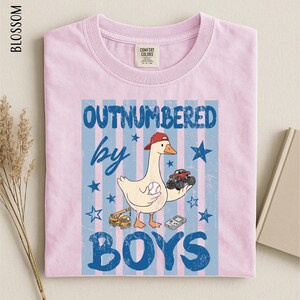 A pink t-shirt with a graphic design featuring a cartoon duck holding a baseball and a toy truck, along with the text "OUTNUMBERED BY BOYS".