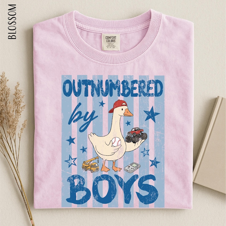 A pink t-shirt with a graphic design featuring a cartoon duck holding a baseball and a toy truck, along with the text "OUTNUMBERED BY BOYS".