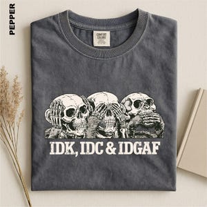 May include: A charcoal gray t-shirt featuring a graphic of three skeletons mimicking the see no evil, hear no evil, speak no evil pose. Below the skeletons, the text reads IDK, IDC & IDGAF in a bold, white, distressed font. The shirt has a crew neck.