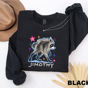 May include: A black long-sleeve sweatshirt featuring a graphic of a raccoon with shaggy fur. The design includes colorful hand-drawn style stars and the name JIMOTHY printed in white capital letters below the animal illustration.
