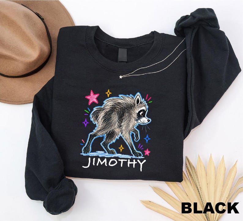 May include: A black long-sleeve sweatshirt featuring a graphic of a raccoon with shaggy fur. The design includes colorful hand-drawn style stars and the name JIMOTHY printed in white capital letters below the animal illustration.