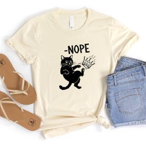 May include: A cream-coloured t-shirt featuring a black cat kicking a crown, with the word "-NOPE" above. The shirt is styled with blue denim shorts and brown sandals.