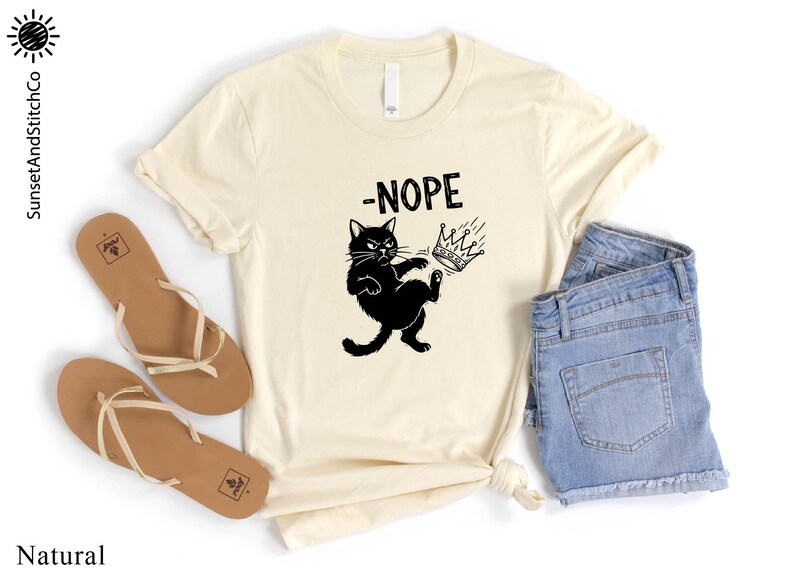 May include: A cream-coloured t-shirt featuring a black cat kicking a crown, with the word "-NOPE" above. The shirt is styled with blue denim shorts and brown sandals.