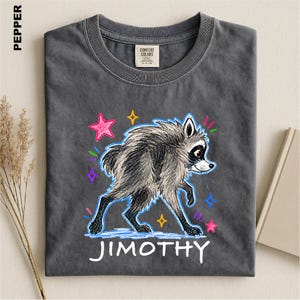 May include: A charcoal gray t-shirt featuring a graphic of a raccoon drawn in a crayon style. The raccoon is surrounded by colorful stars and small marks. The name JIMOTHY is printed in white capital letters below the raccoon illustration.