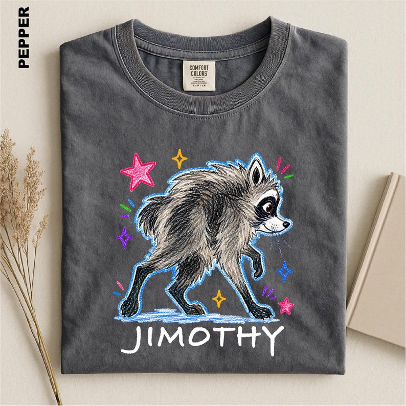 May include: A charcoal gray t-shirt featuring a graphic of a raccoon drawn in a crayon style. The raccoon is surrounded by colorful stars and small marks. The name JIMOTHY is printed in white capital letters below the raccoon illustration.