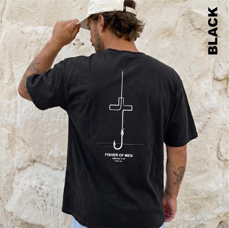 May include: A black short-sleeve t-shirt featuring a white line art graphic on the back. The design depicts a fishing hook shaped like a cross with the text "FISHER OF MEN", "Matthew 4:19", and "Follow me" printed underneath the graphic.