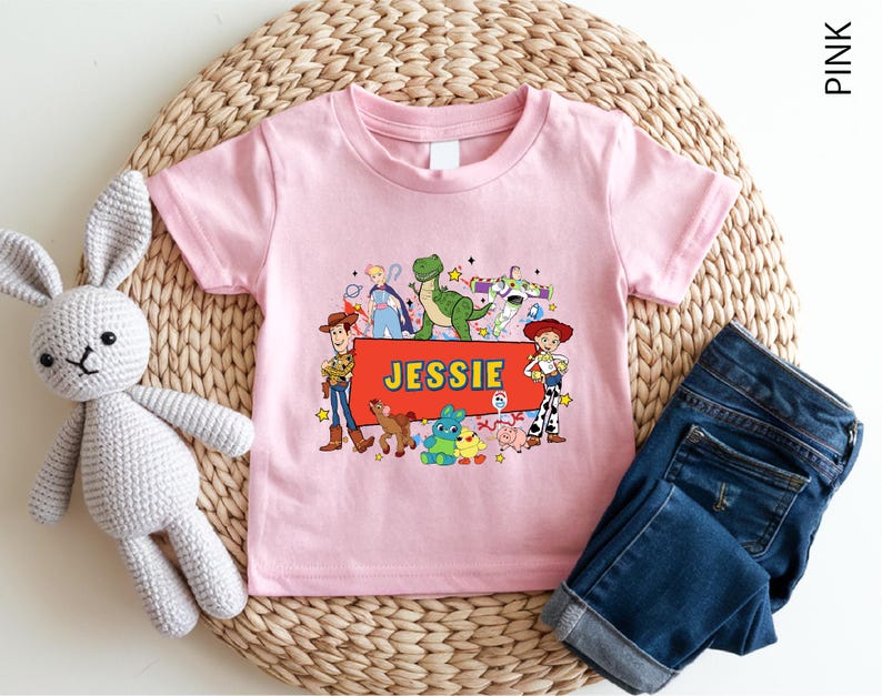 A pink t-shirt with a cartoon character design and the name "JESSIE" printed on it, along with a crocheted rabbit toy and a pair of blue jeans.