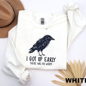May include: A white long-sleeve sweatshirt featuring a black graphic illustration of a crow. Below the bird, the text reads, "I GOT UP EARLY THERE WAS NO WORM" in a bold, black sans-serif font. The garment has a crew neckline and ribbed cuffs.