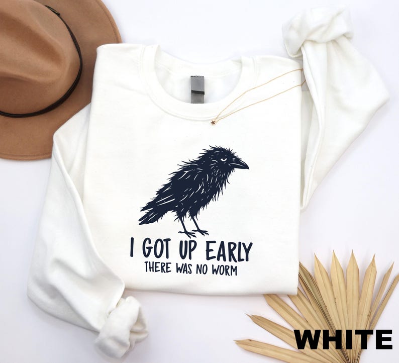 May include: A white long-sleeve sweatshirt featuring a black graphic illustration of a crow. Below the bird, the text reads, "I GOT UP EARLY THERE WAS NO WORM" in a bold, black sans-serif font. The garment has a crew neckline and ribbed cuffs.