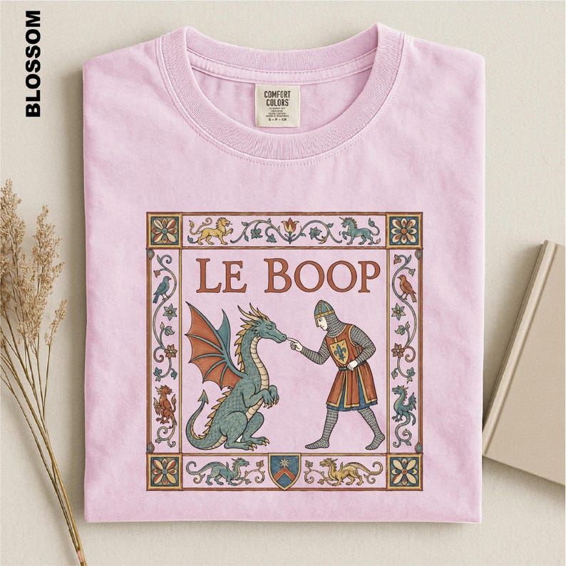 May include: A light pink t-shirt featuring a medieval-style graphic print. The design shows a knight touching a dragon's nose with the text LE BOOP centered above. The illustration is framed by a decorative border with small mythical creatures.