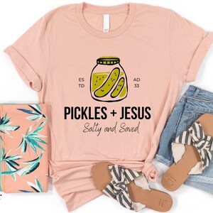 a pink t-shirt with a graphic design, a pair of blue jeans, a pink and green floral bag, and a pair of black and white striped sandals, all laid out on a white background.