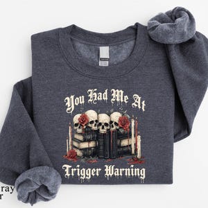 May include: Dark gray heather sweatshirt with the text "You Had Me At Trigger Warning." The graphic features skulls, roses, books, and candles. The sweatshirt has a crew neck and long sleeves.
