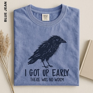 May include: A blue jean colored short-sleeve t-shirt featuring a dark navy graphic of a ruffled crow. Below the bird, the text reads: I GOT UP EARLY THERE WAS NO WORM. The shirt has a crew neckline and a Comfort Colors brand label at the inner neck.