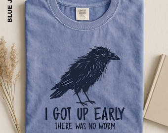 Puede incluir: Una camiseta de manga corta color azul vaquero con un gráfico azul marino de un cuervo despeinado. Debajo del ave, el texto dice: I GOT UP EARLY THERE WAS NO WORM. La camiseta tiene cuello redondo y una etiqueta de la marca Comfort Colors.