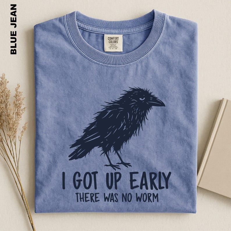 May include: A blue jean colored short-sleeve t-shirt featuring a dark navy graphic of a ruffled crow. Below the bird, the text reads: I GOT UP EARLY THERE WAS NO WORM. The shirt has a crew neckline and a Comfort Colors brand label at the inner neck.
