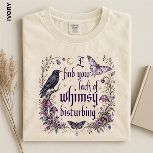 Puede incluir: Una camiseta color marfil con un estampado gr&aacute;fico que muestra un cuervo negro, mariposas moradas, setas y enredaderas florales. El texto dice, I find your lack of whimsy disturbing. El dise&ntilde;o incluye peque&ntilde;as estrellas y una luna creciente en tinta morada.