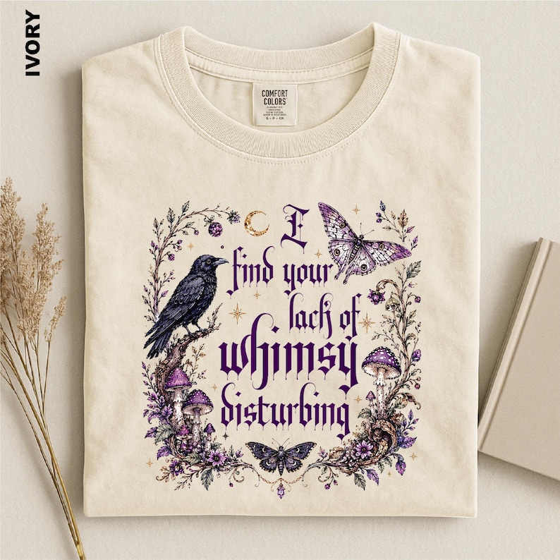Puede incluir: Una camiseta color marfil con un estampado gr&aacute;fico que muestra un cuervo negro, mariposas moradas, setas y enredaderas florales. El texto dice, I find your lack of whimsy disturbing. El dise&ntilde;o incluye peque&ntilde;as estrellas y una luna creciente en tinta morada.