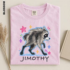 May include: A light pink t-shirt featuring a graphic of a gray and black raccoon with a sketch-like texture. The raccoon is surrounded by colorful hand-drawn stars and sparkles. The name JIMOTHY is printed in black capital letters below the raccoon.