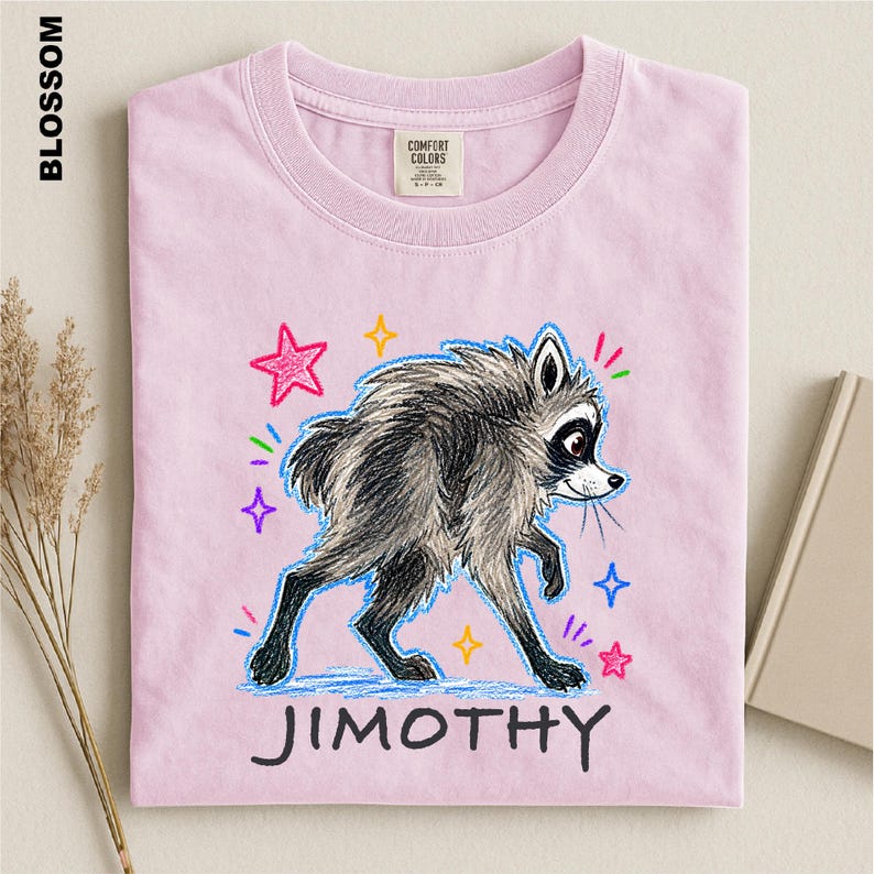 May include: A light pink t-shirt featuring a graphic of a gray and black raccoon with a sketch-like texture. The raccoon is surrounded by colorful hand-drawn stars and sparkles. The name JIMOTHY is printed in black capital letters below the raccoon.