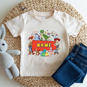A white t-shirt with a colorful graphic of characters from the Toy Story movie, including Woody, Buzz Lightyear, and other beloved characters, along with a name tag. The t-shirt is placed on a woven placemat, and there is a crocheted stuffed rabbit next to it.