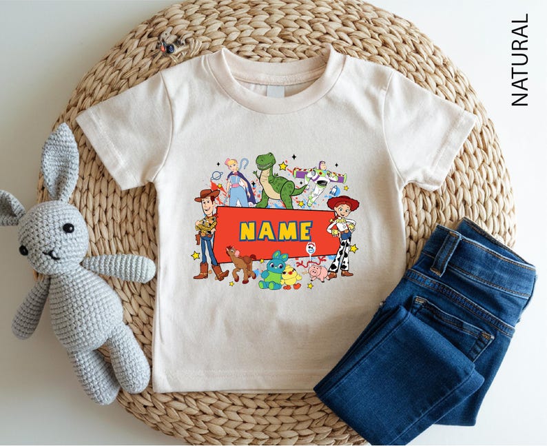 A white t-shirt with a colorful graphic of characters from the Toy Story movie, including Woody, Buzz Lightyear, and other beloved characters, along with a name tag. The t-shirt is placed on a woven placemat, and there is a crocheted stuffed rabbit next to it.