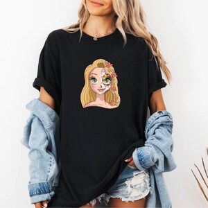 A woman wearing a black t-shirt with a cartoon illustration of a blonde woman with a flower in her hair. She is also wearing a denim jacket and denim shorts.